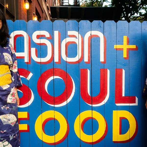 Two smiling women in traditional kimonos pose next to a vibrant "ASIAN + SOUL FOOD" sign.