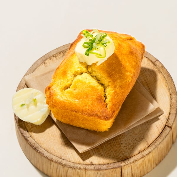 Golden cornbread loaf with butter and chives on a wooden plate with a side of butter.