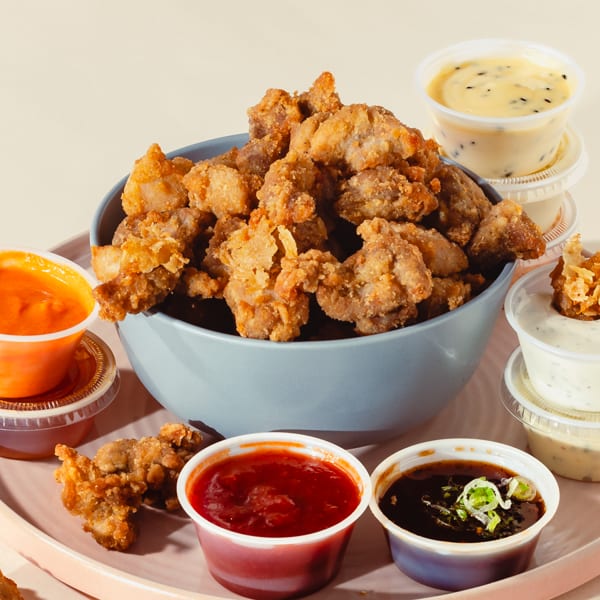 A blue bowl of crispy fried chicken bites surrounded by various dipping sauces on a pink tray.