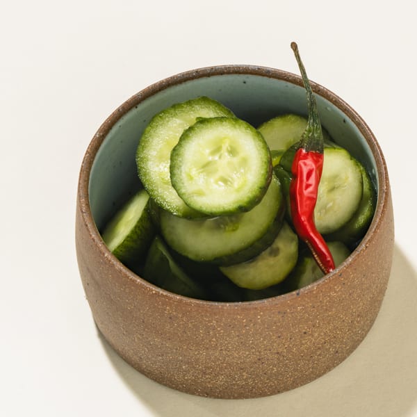 A brown bowl filled with sliced cucumbers and a single red chili pepper on a white surface.