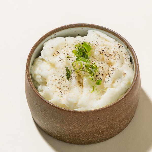 A rustic brown bowl filled with creamy mashed potatoes, topped with black pepper and chopped green onions.